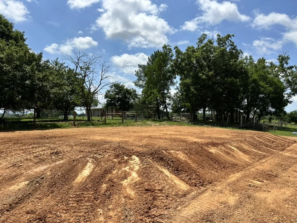 A dirt field with tire tracks, surrounded by trees and a wooden fence under a partly cloudy sky.