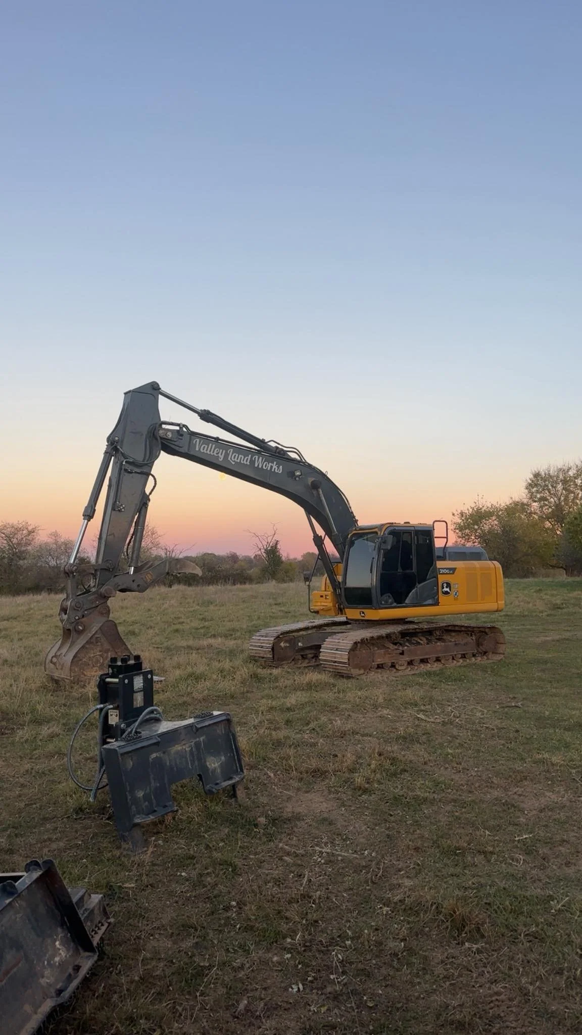A yellow and black excavator with the words 'Valley Land Works' on the arm, positioned on a grassy field at sunset or sunrise, with some equipment in the foreground.