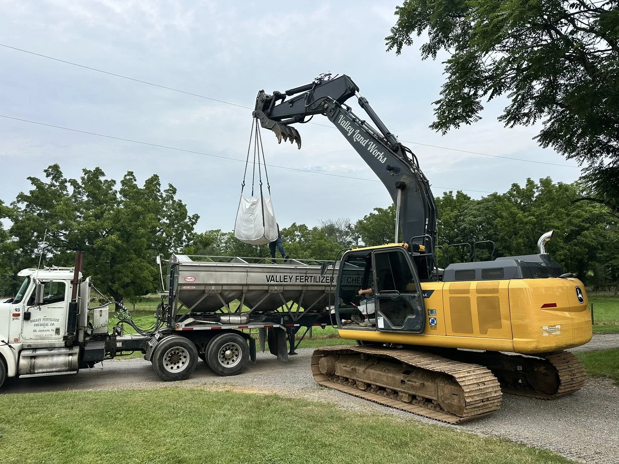 A construction site with a large excavator lifting a bag, a fertilizer truck, and a generator, surrounded by trees and a cloudy sky.