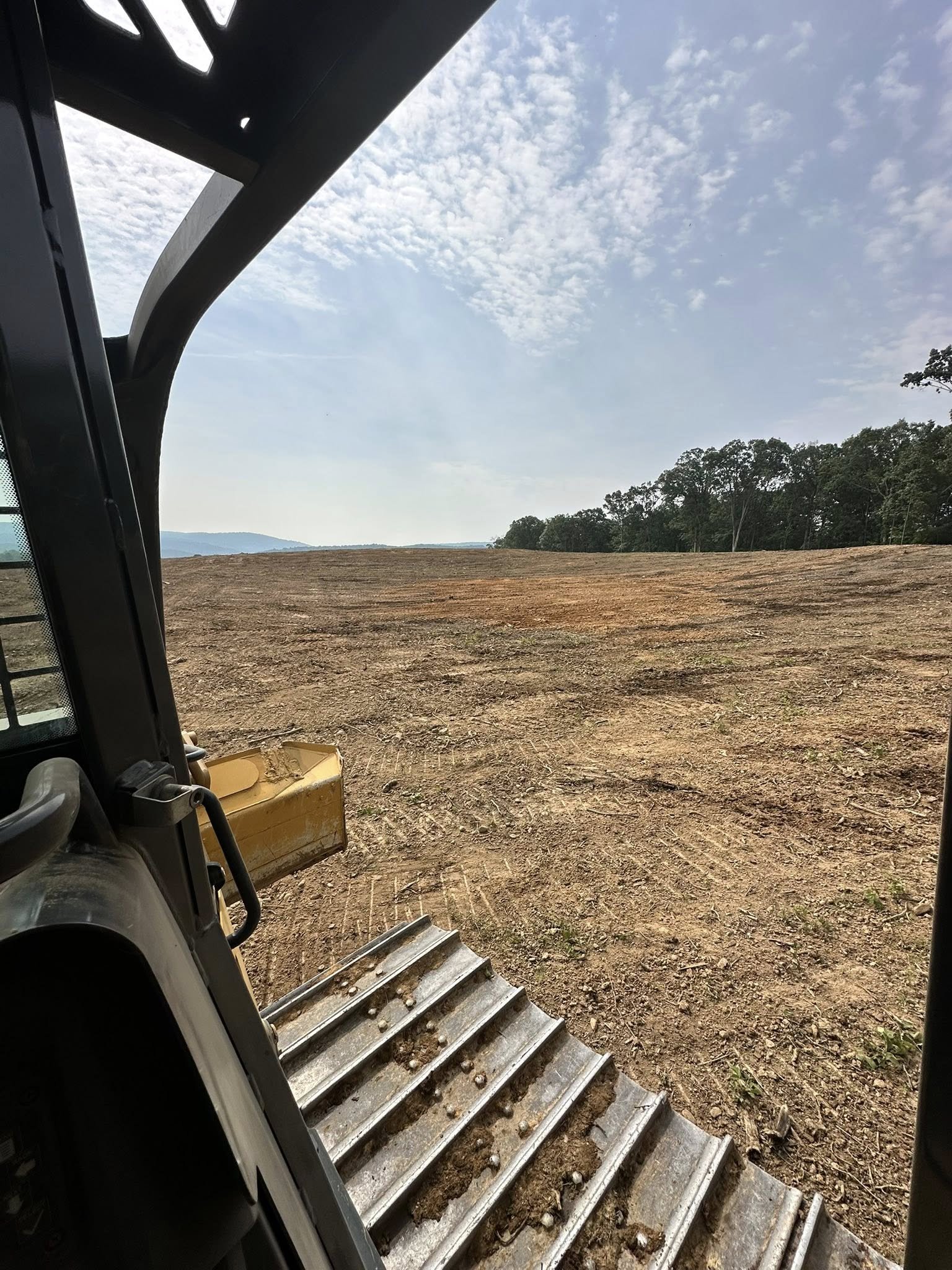 View from inside construction vehicle looking out over a cleared, dirt-covered construction site with trees and hills in the distance under a partly cloudy sky.