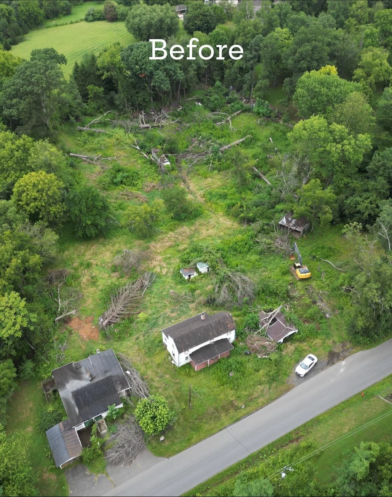 An aerial view of a residential area with several houses, trees, and a large open green space where fallen trees are visible. A yellow excavator is working near the fallen trees.