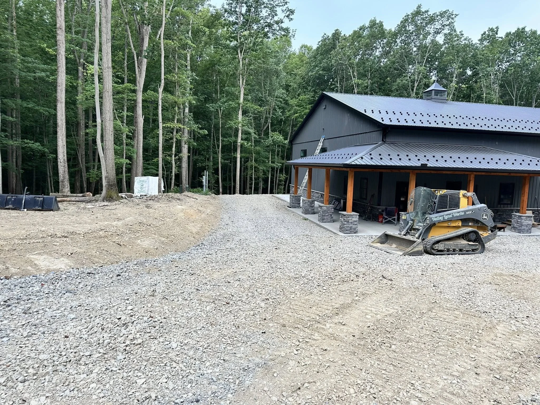 Construction site with gravel ground, partially built house with metal roof, surrounded by trees, construction equipment including a small excavator, ladder, and building materials.