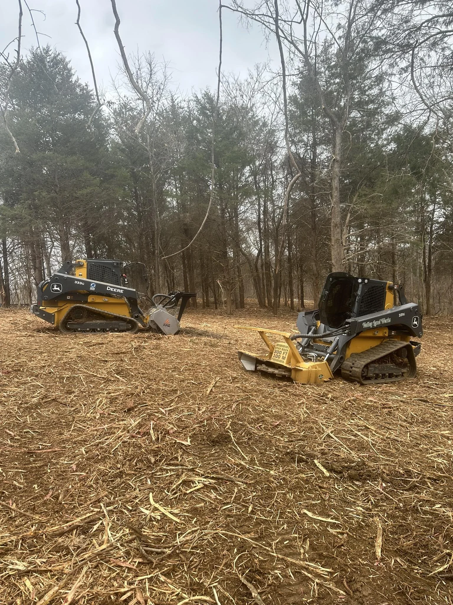 Two small yellow bulldozer machines on dirt ground in a wooded area with tall, leafless and evergreen trees in the background.