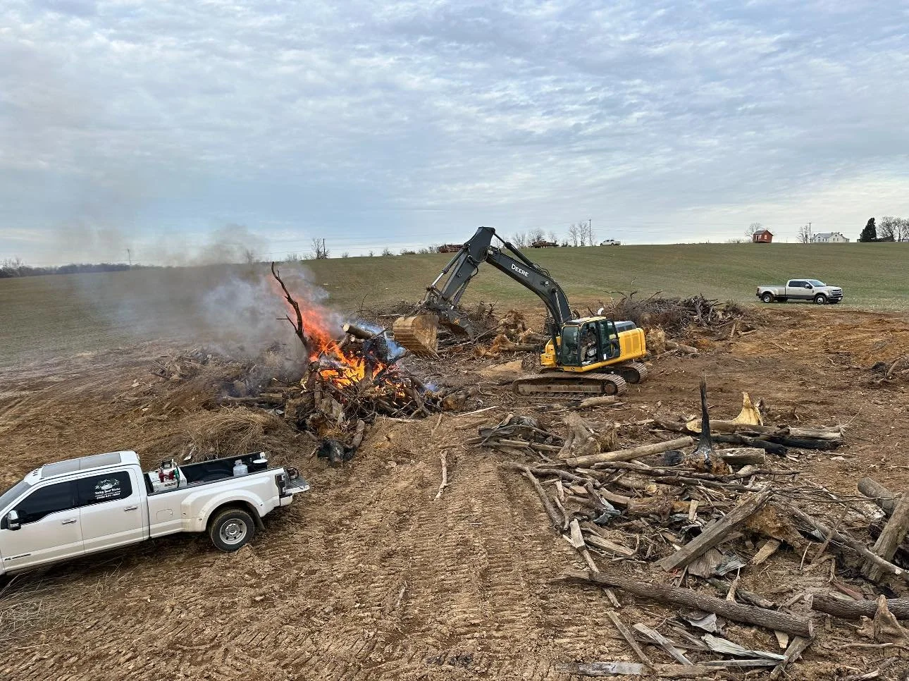 A construction site with an excavator clearing debris among burning and smoldering wood, with pickup trucks parked on dirt and a grassy hill with a few houses in the background.