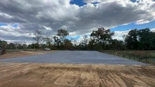 Wide view of an empty gravel lot with trees and cloudy sky in the background.