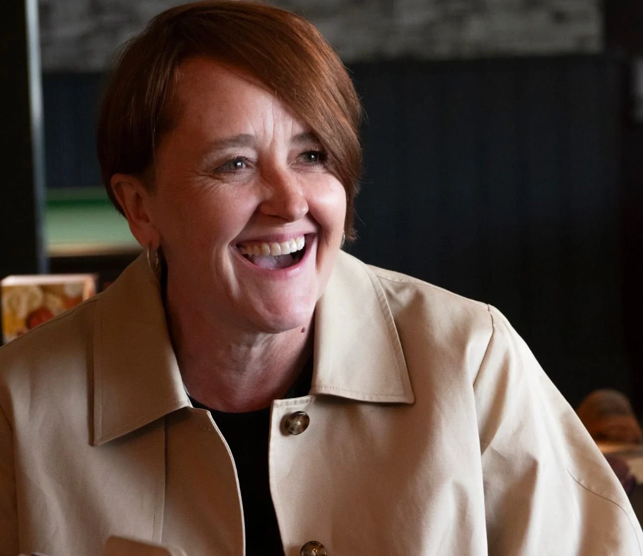 A woman with short red hair smiling and laughing, wearing a beige jacket in an indoor setting.
