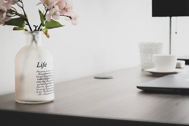 A white vase with pink flowers and black text that says "Life" on a wooden surface, with a computer monitor, a white cup and saucer, and a glass container in the background.