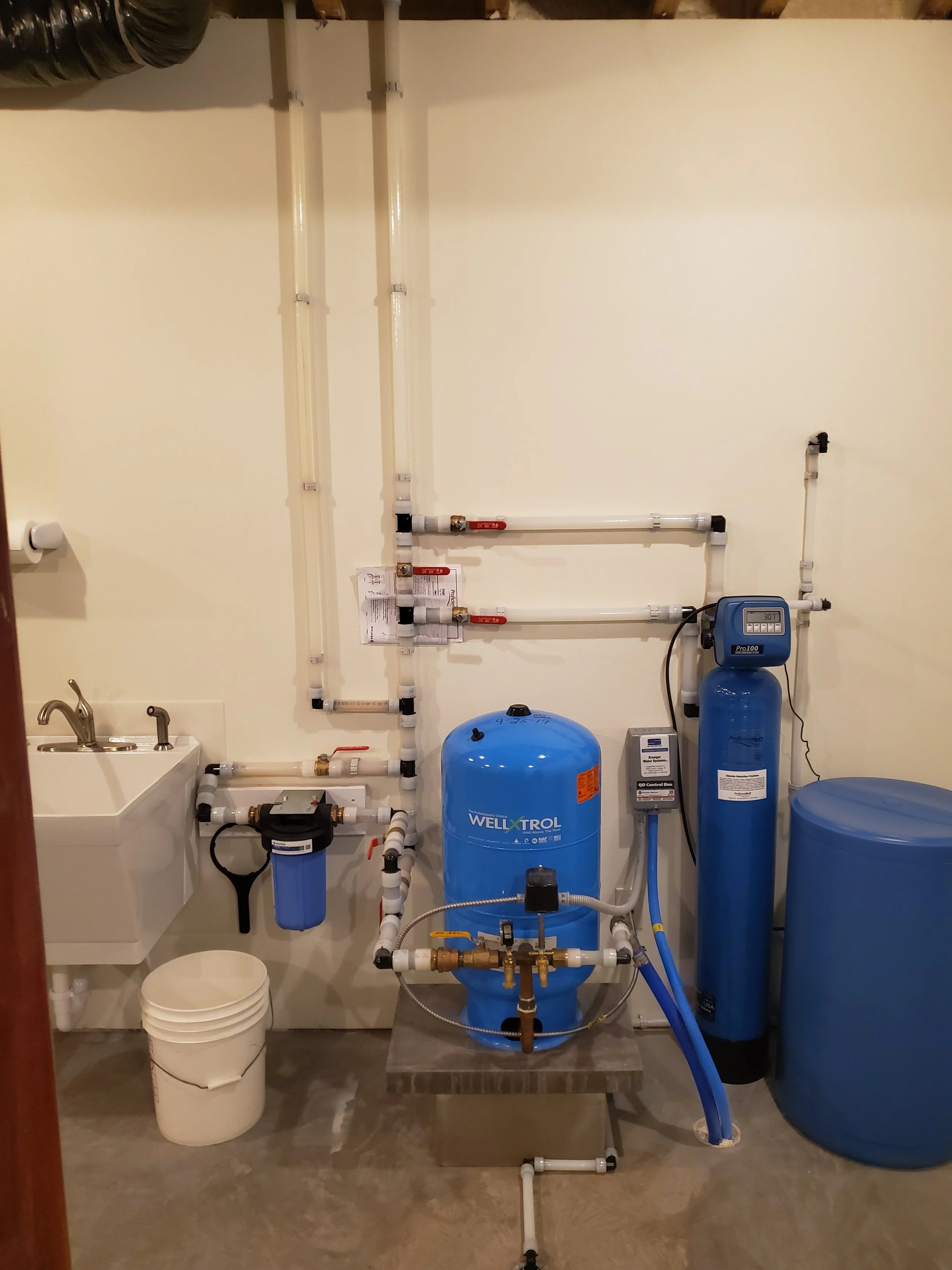 A well tankless water filtration system with blue tanks, pipes, and gauges installed in a utility room.