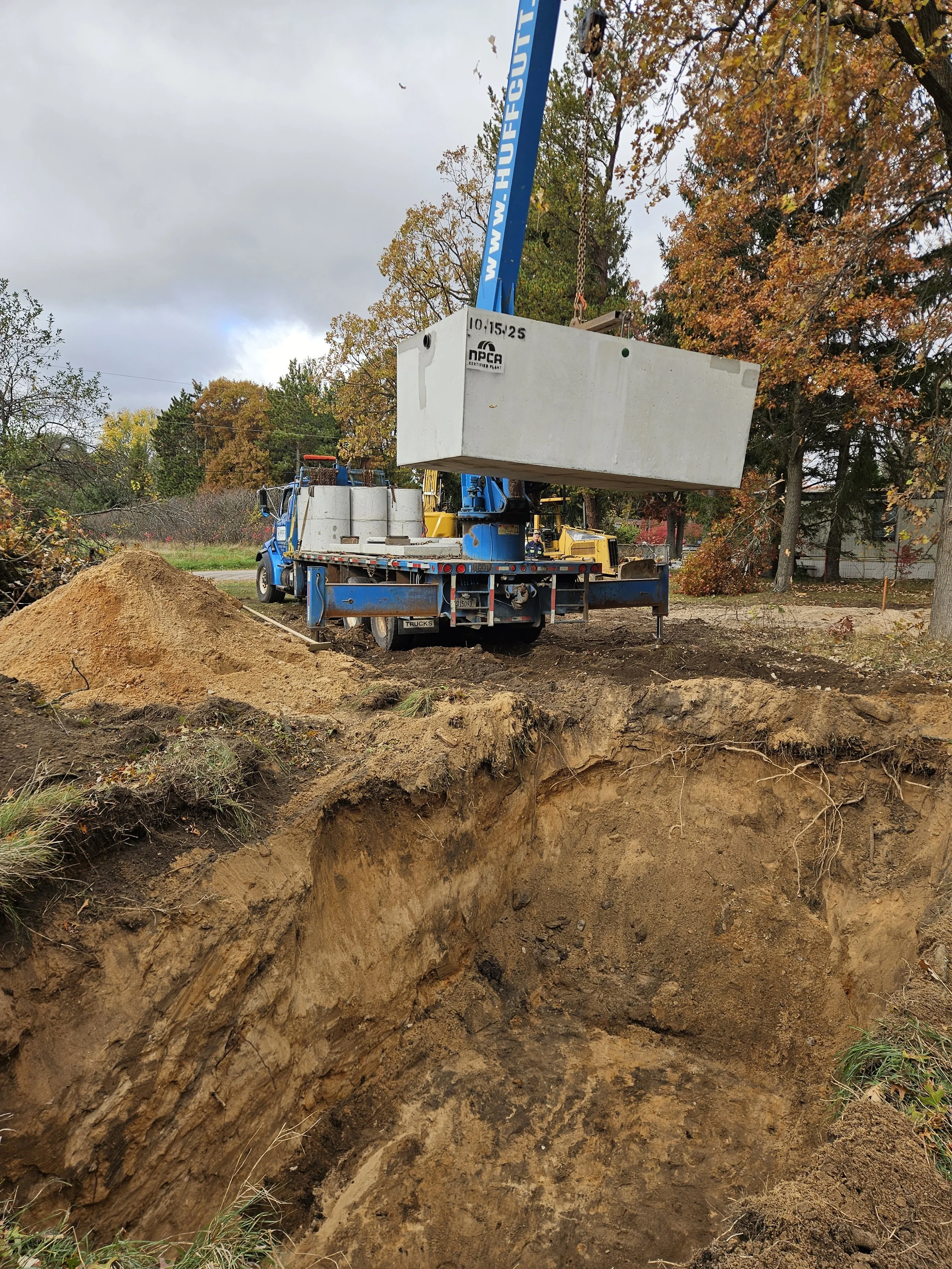 Construction site with a large truck and crane lifting a concrete block over a deep excavated hole in the ground, surrounded by trees with autumn foliage.