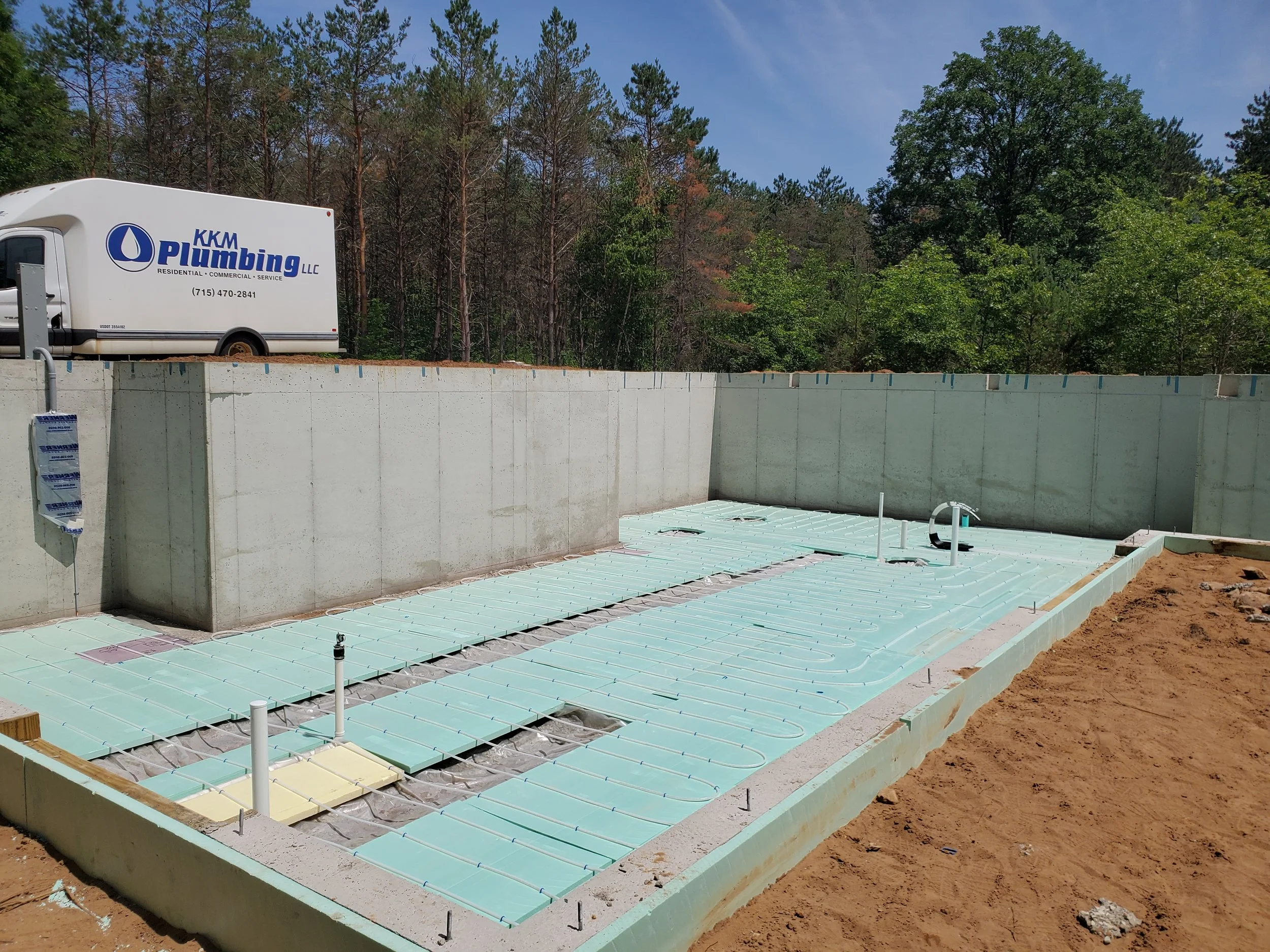 Construction site with pool plumbing and insulation, concrete walls, and a KKM Plumbing LLC truck parked on the partially built structure, surrounded by trees and clear blue sky.