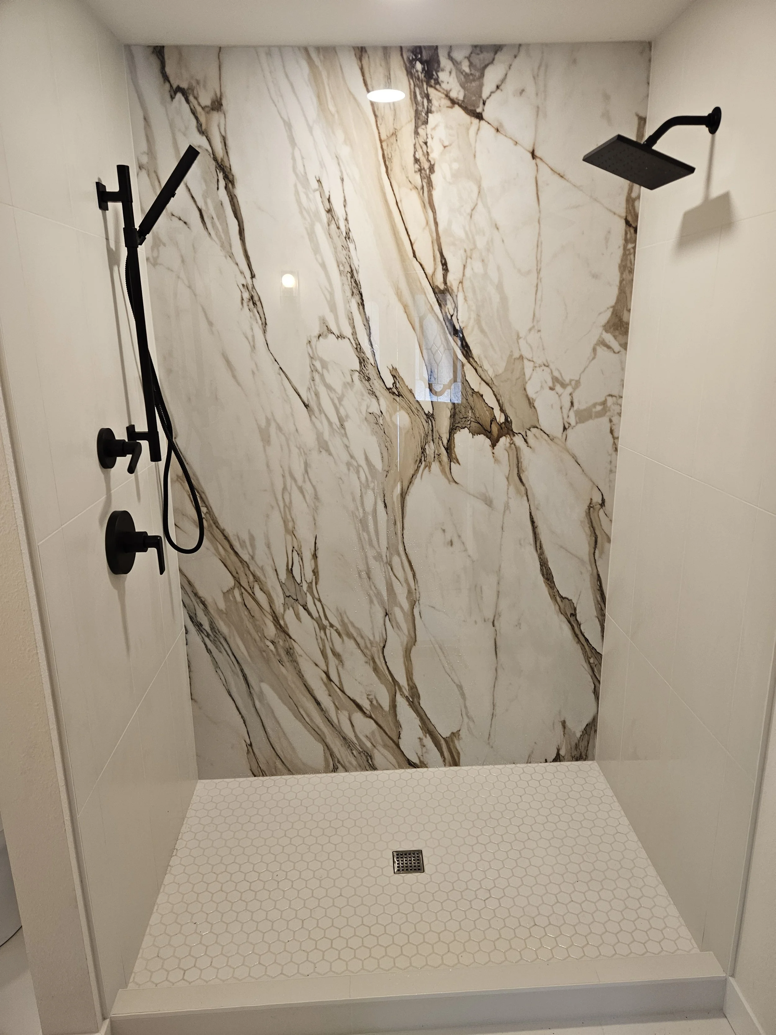 A modern walk-in shower with a large, marble-patterned wall and a white hexagonal tile floor. It has a black rainfall showerhead on the ceiling and a black wall-mounted shower panel on the left side with additional controls and a hand-held shower attachment.