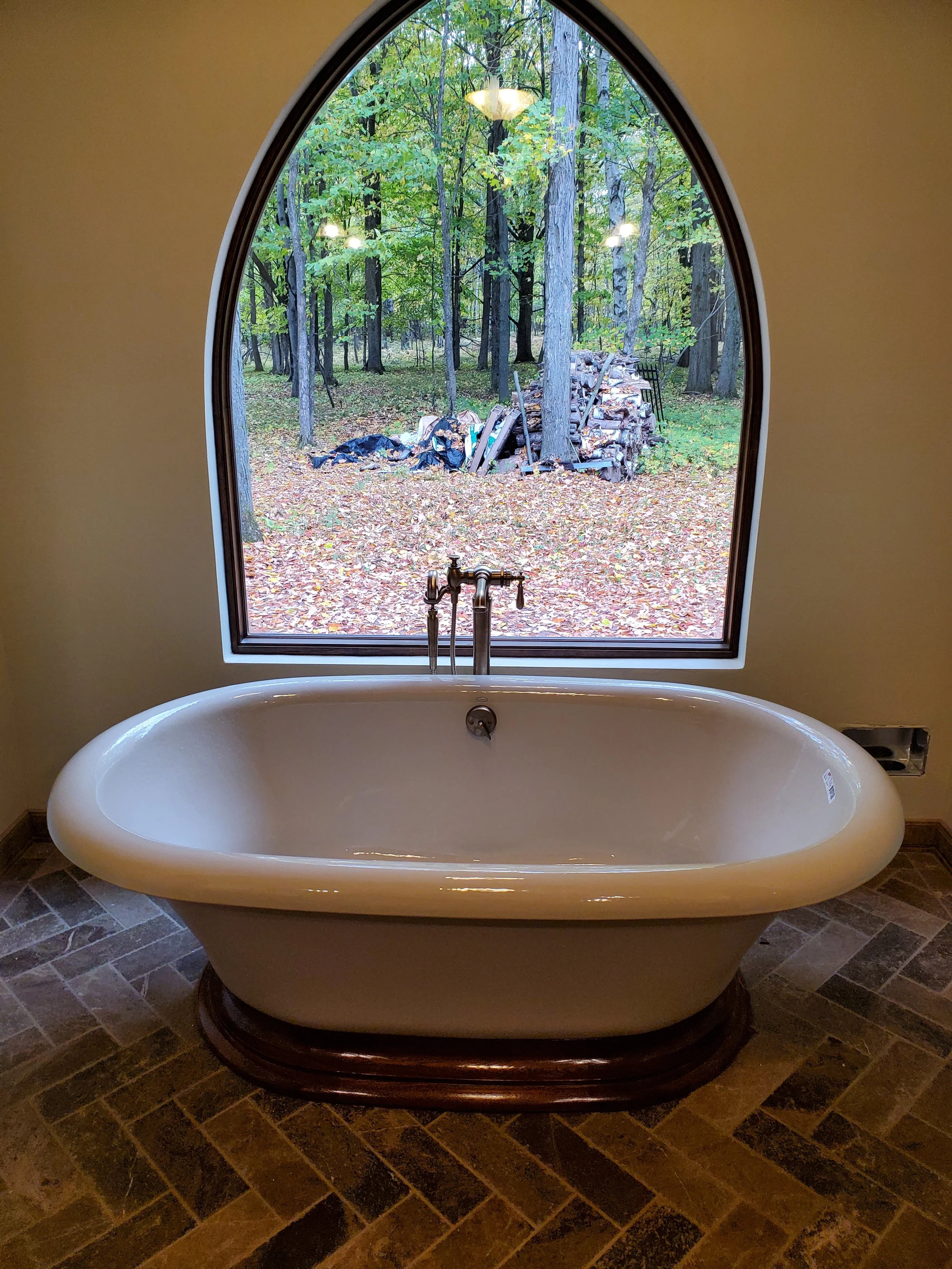 Free-standing bathtub with dark wood base in front of large arched window looking out onto a wooded forest scene with trees and fallen leaves.
