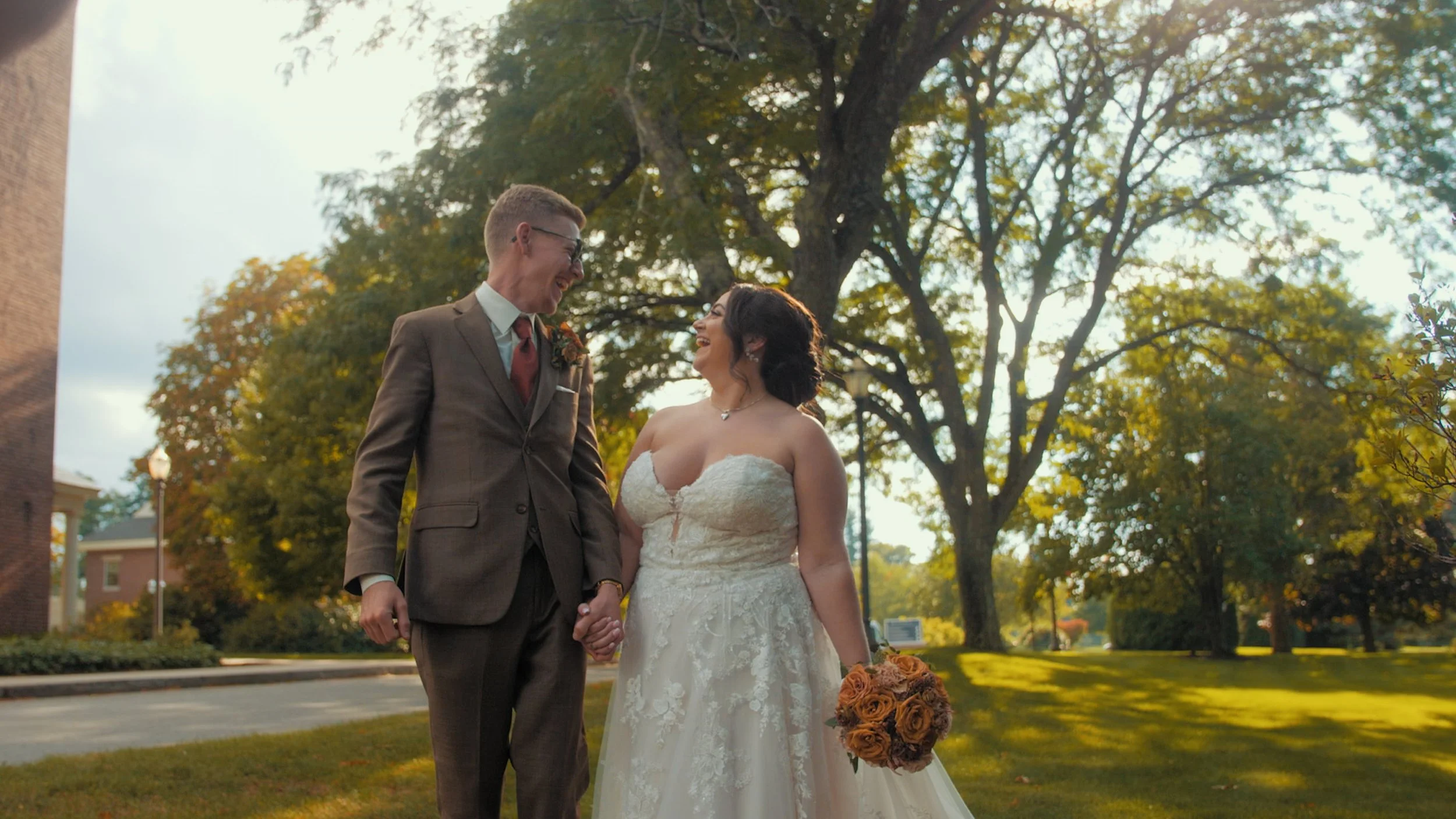 A newlywed couple holding hands and smiling at each other outdoors during daytime, with trees and a brick building in the background.