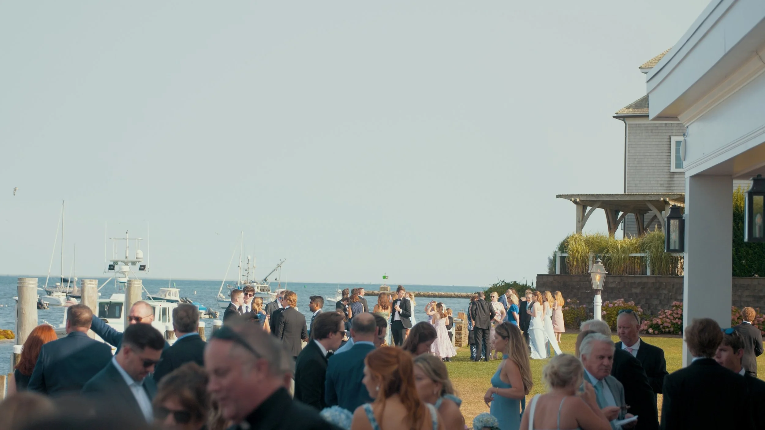 People dressed in formal attire gathering at a waterfront outdoor event near boats and yachts, with a building and a clear sky in the background.