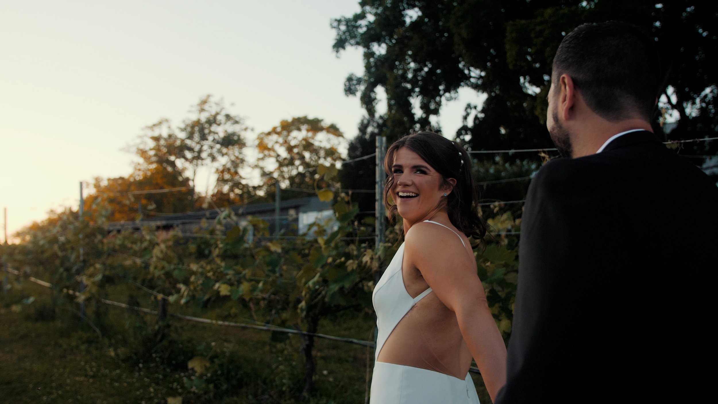 A bride and groom holding hands outdoors during sunset, with the bride smiling and looking back at the groom.