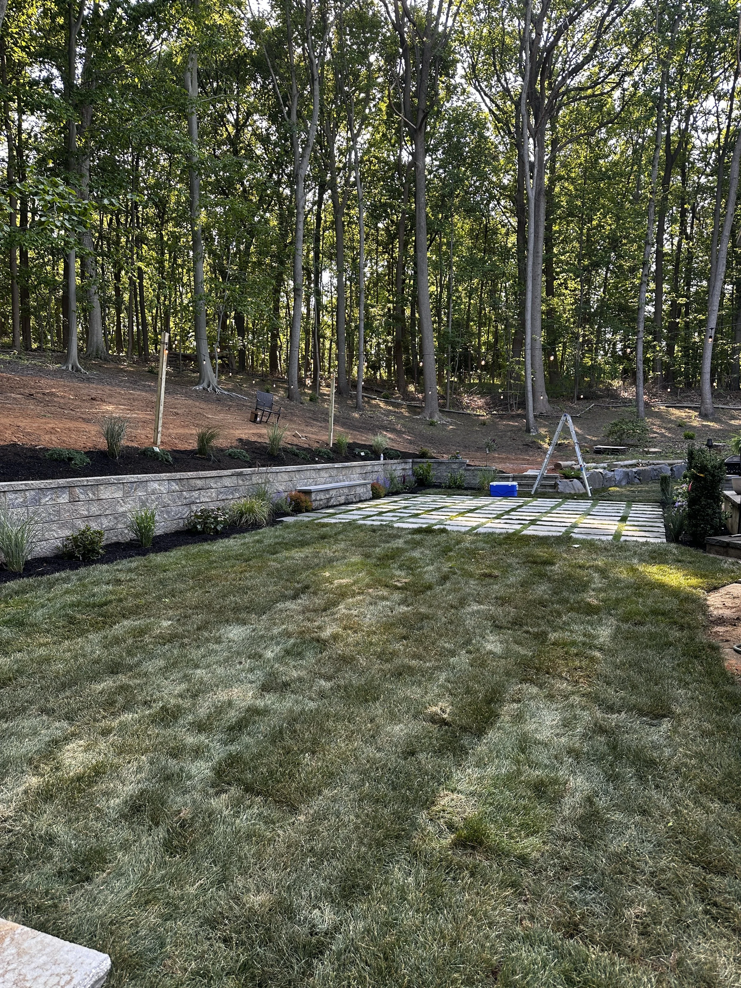 Backyard with a grassy lawn, a stone patio with grid-like pattern, a small swing set, and a wooded hillside in the background.