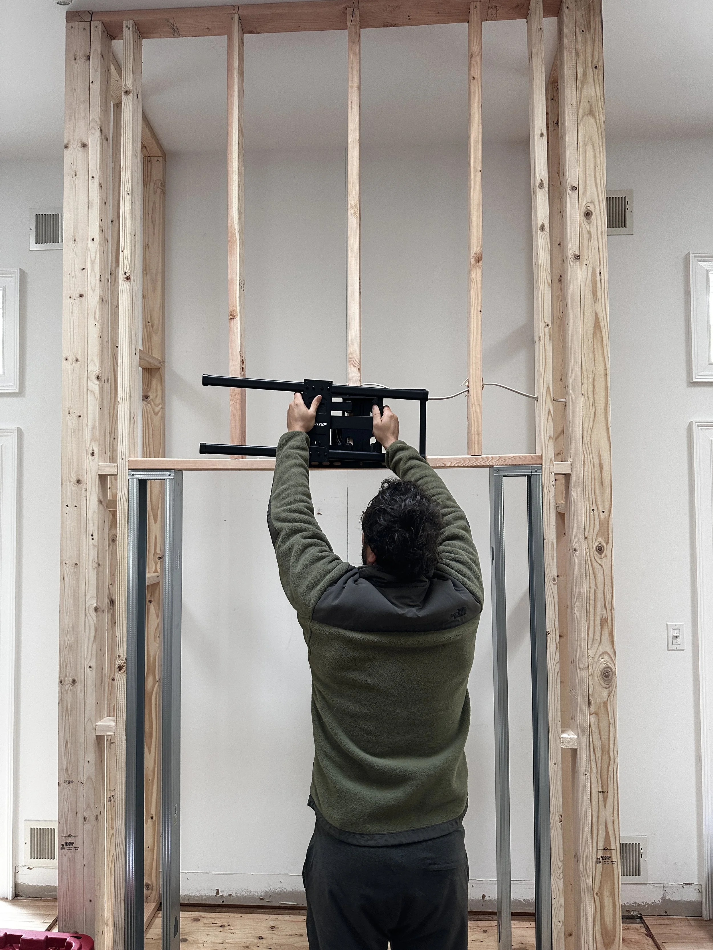 A man installing or working on a wooden wall frame in a room. He is holding a black tool, possibly a level or measurement device, and is facing the wall structure while standing on the floor.