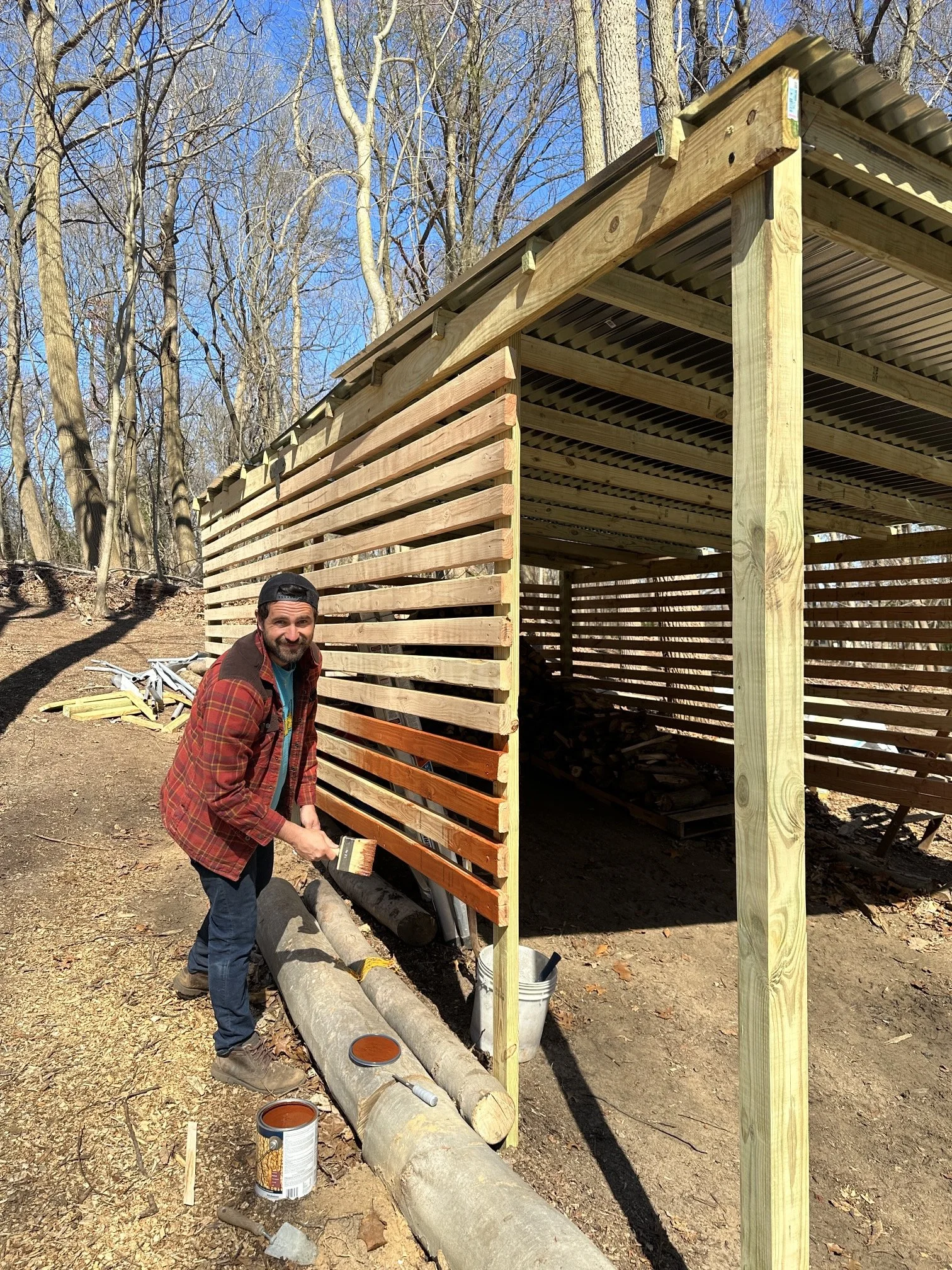 A man painting horizontal wooden slats on a small outdoor wooden structure in a wooded area, with trees and blue sky in the background.