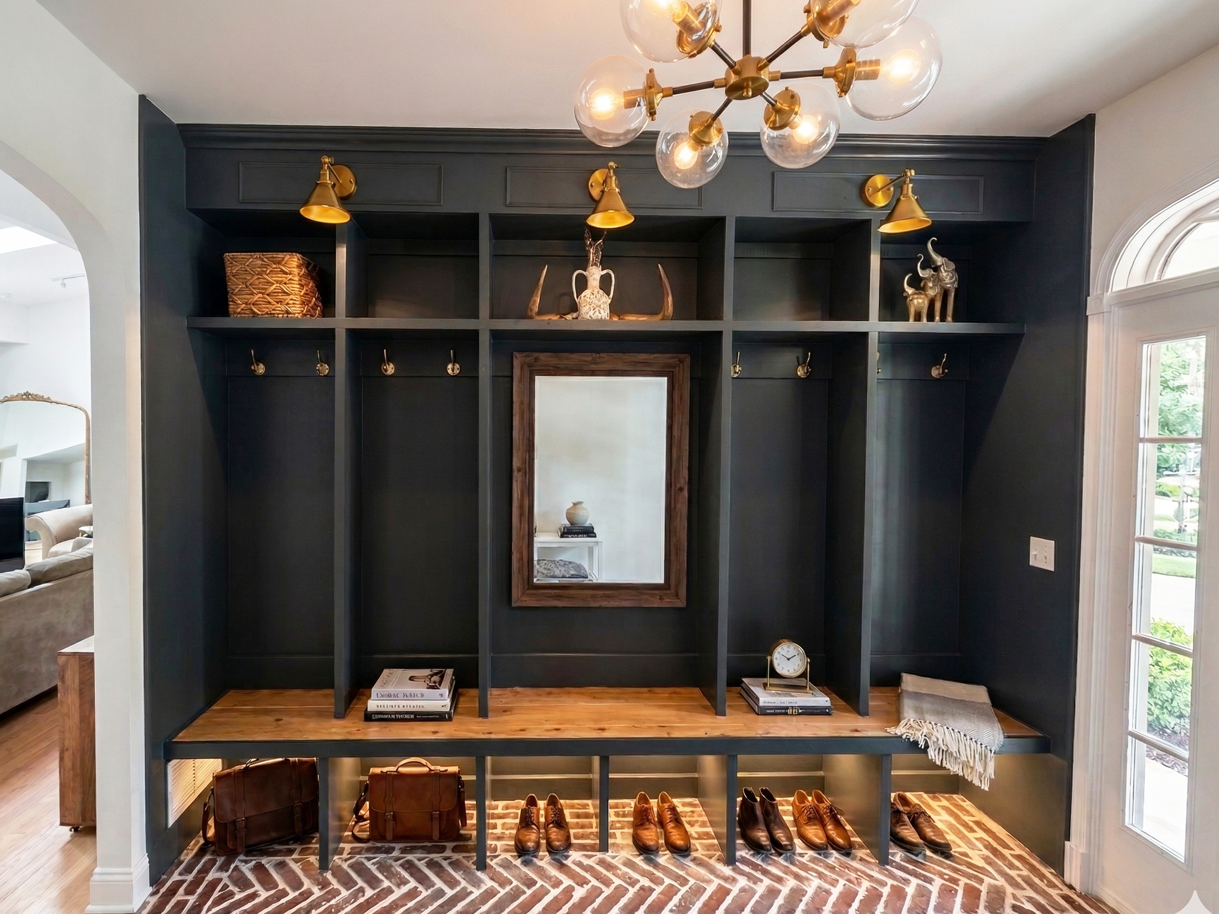 Entryway mudroom with black built-in counter, shelves, and hooks, decorated with books, a mirror, clock, and shoes, with a brick floor and a large window.