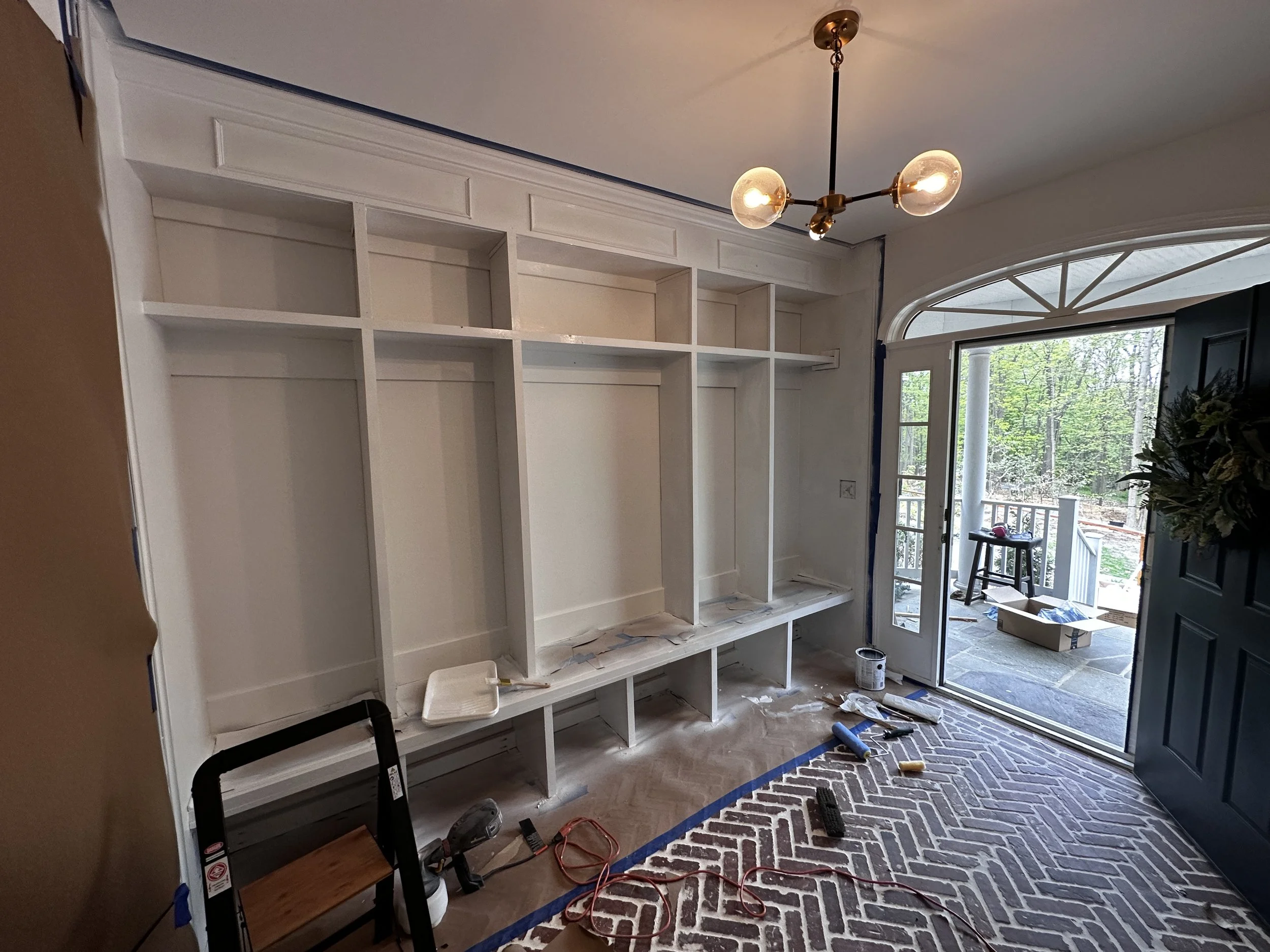 A room under renovation with an empty white built-in bookshelf on the wall, a ladder, construction tools, and painting supplies on the floor. A glass door leads outside to a deck with trees in the background. The room has a hanging light fixture with