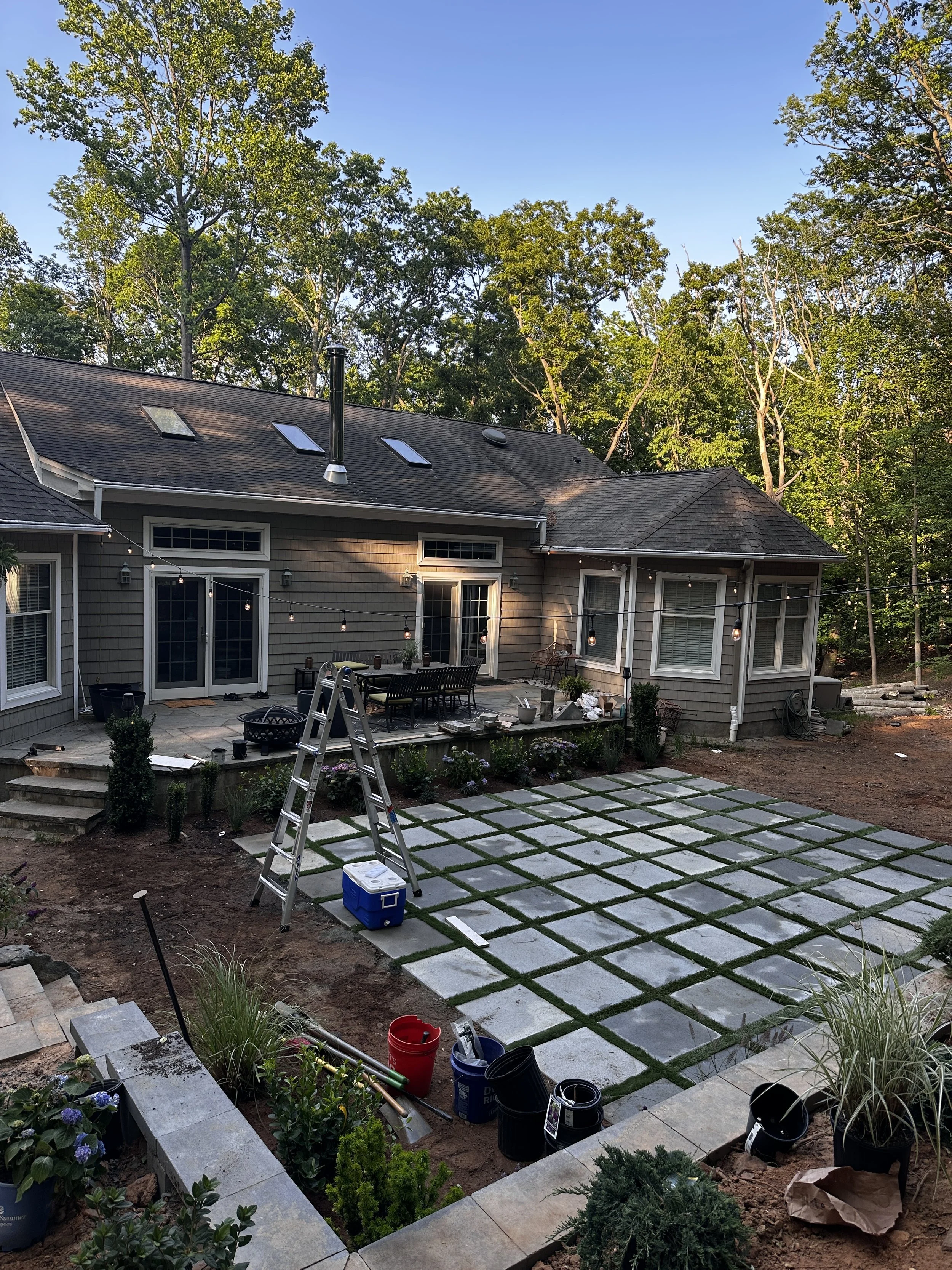 Backyard patio area with paver stones and a house in the background surrounded by trees, construction tools, and furniture.