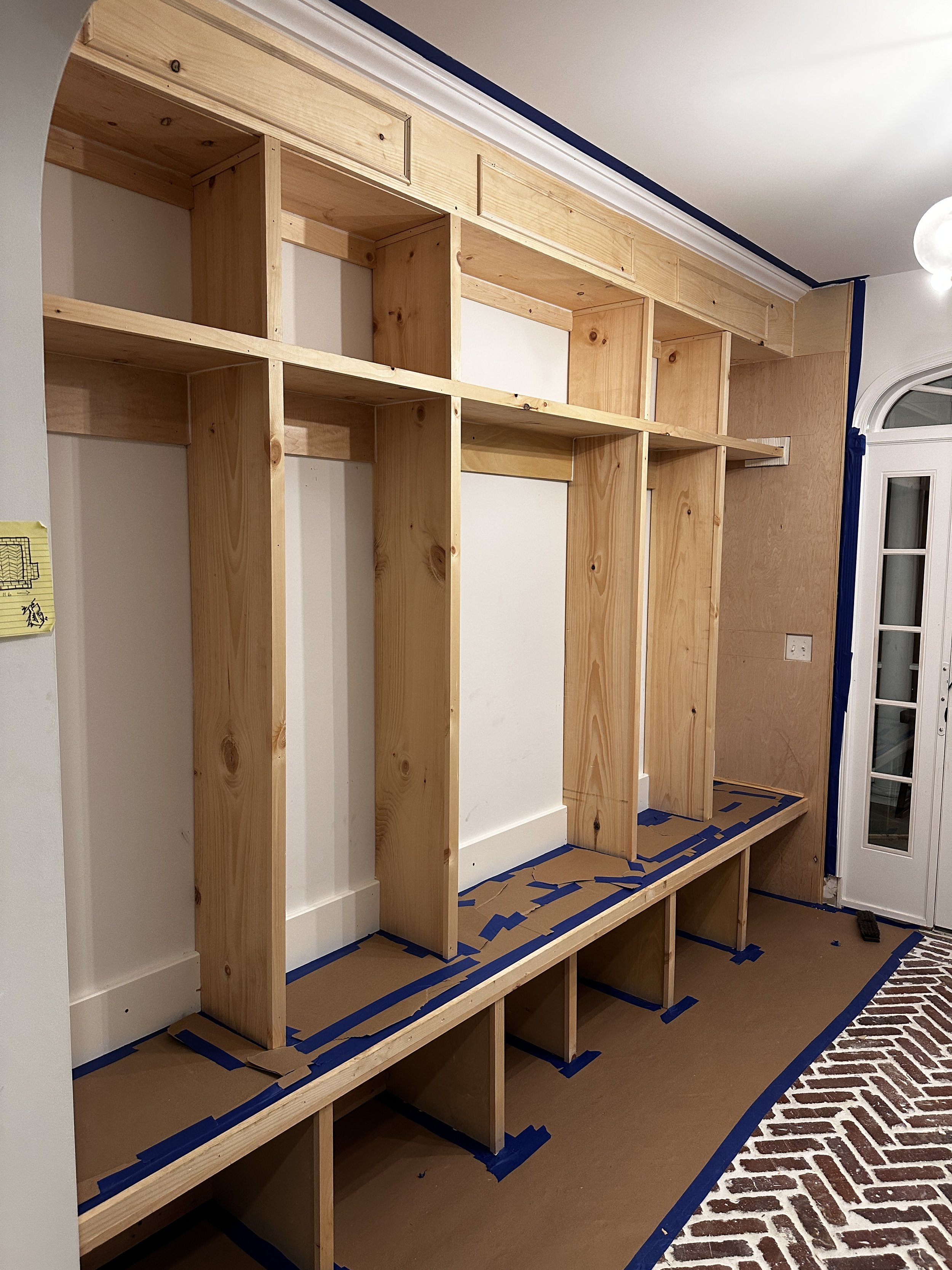 Interior view of a home during construction, showing a built-in wooden shelving unit with open sections, protected floor covered with cardboard and painter's tape, next to a door with glass panels.