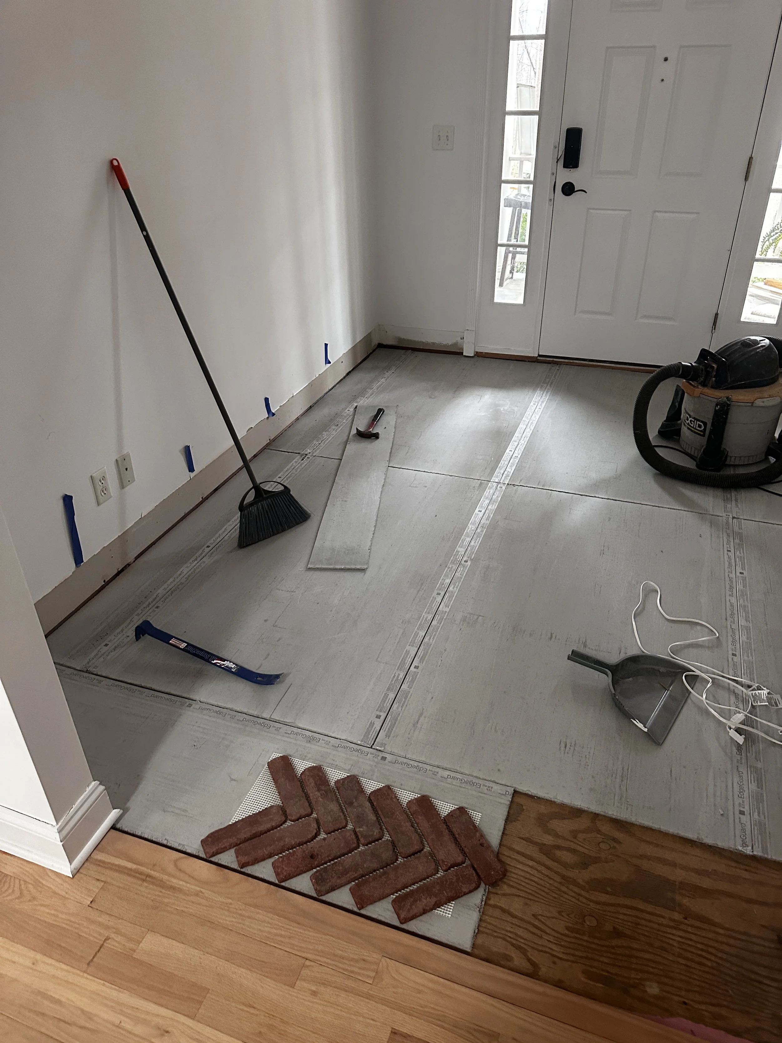 Room under renovation with tools and materials for flooring installation, including brick tiles, a vacuum, a trowel, a saw, and a broom, with partially tiled floor and a door with glass panels.