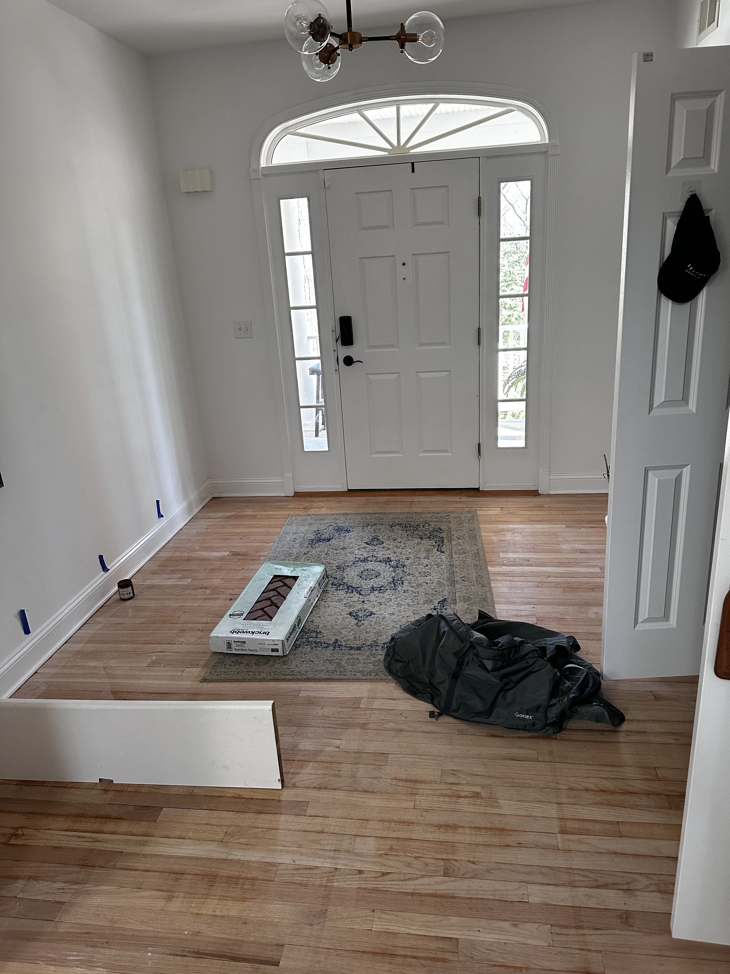 Empty foyer with hardwood floors, white walls, front door with glass sidelights, and a modern chandelier, with items including a black duffel bag, a box, and blue painter's tape on the floor.