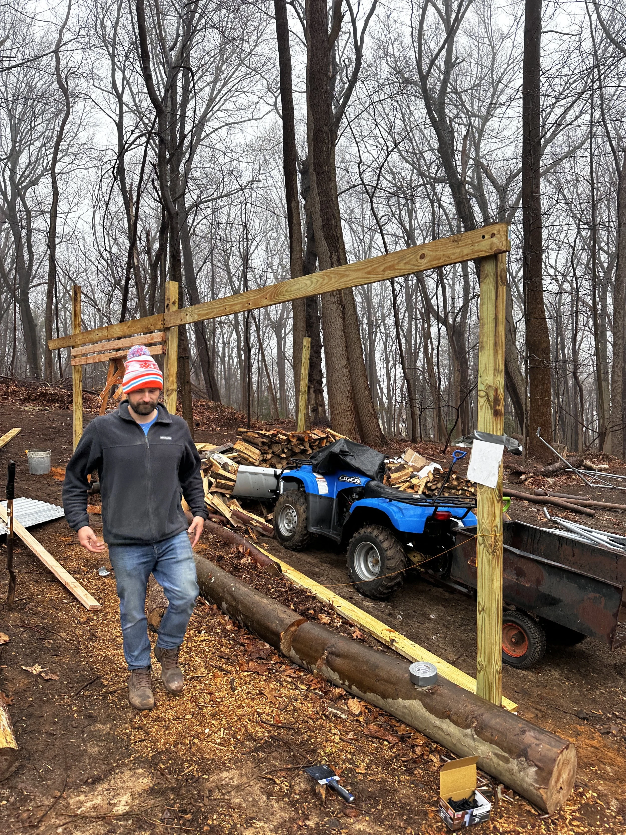 A man in a gray fleece jacket, jeans, and a winter hat with a pom-pom walks in a wooded area under construction. There are stacked logs, a blue utility vehicle, and wooden beams being used for a building project, with trees and a cloudy sky in the ba