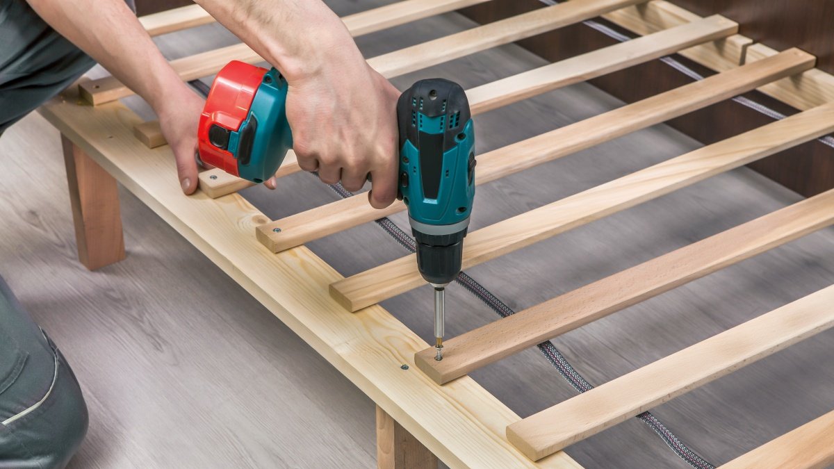 Person using a cordless drill to assemble a wooden bed frame with slats.
