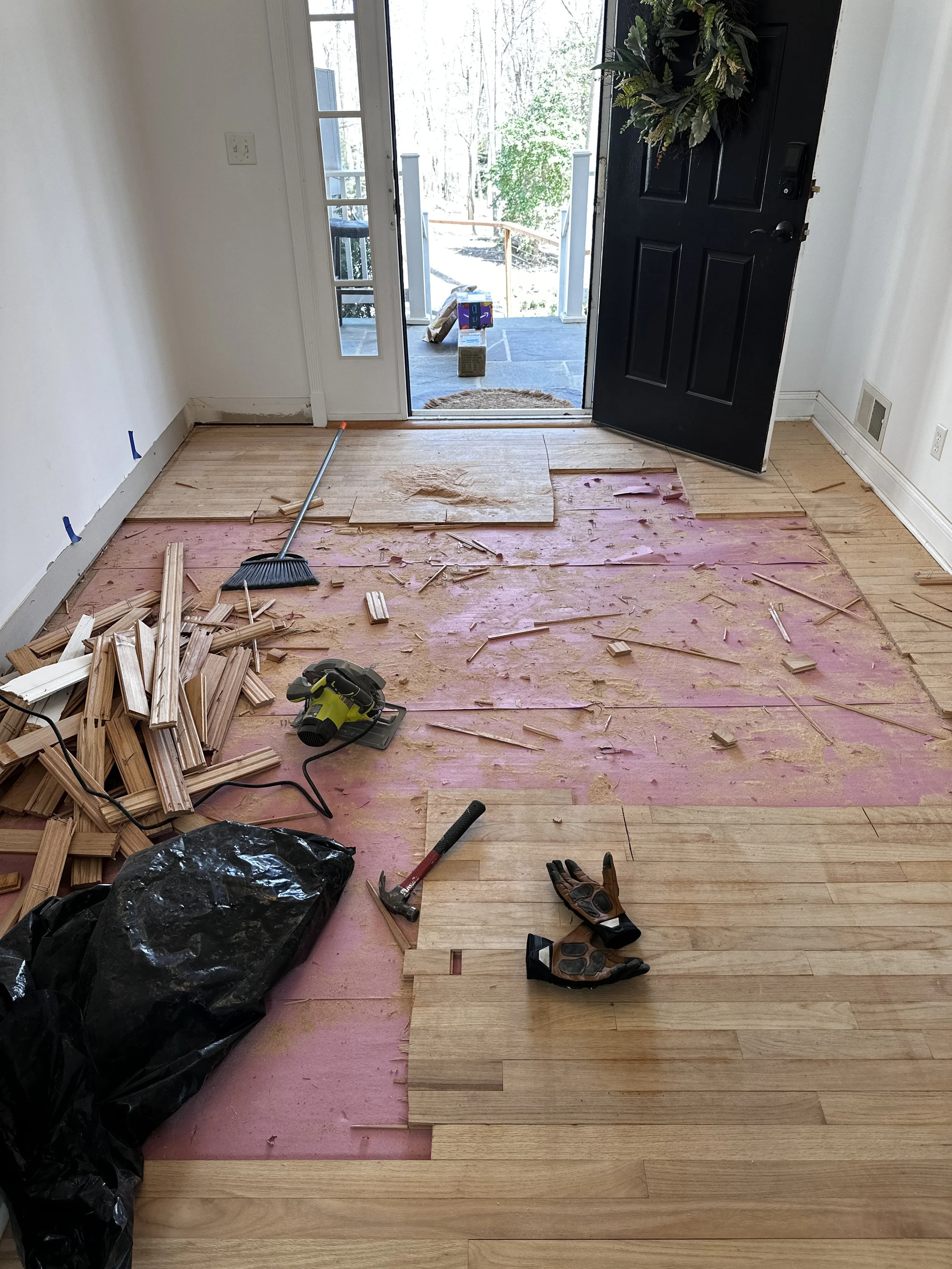Room undergoing hardwood floor installation, with some hardwood planks already laid, tools, and materials scattered around, and an open door leading outside.