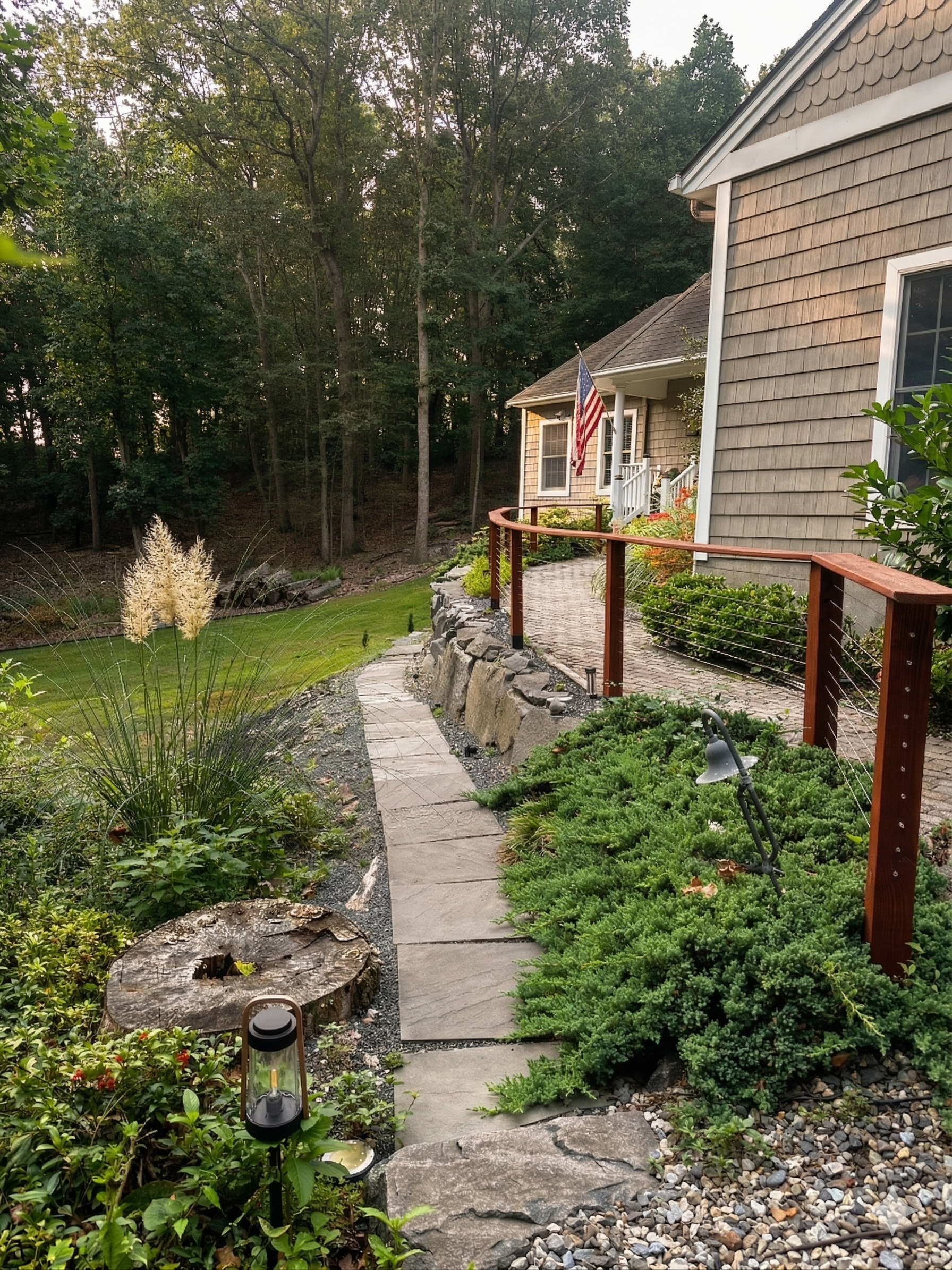 A landscaped backyard with a winding stone pathway leading to a house, bordered by lush green plants, a wooden and wire fence, a tree stump, and garden lights. The house has beige siding, white trim, and an American flag hanging near the entrance, wi