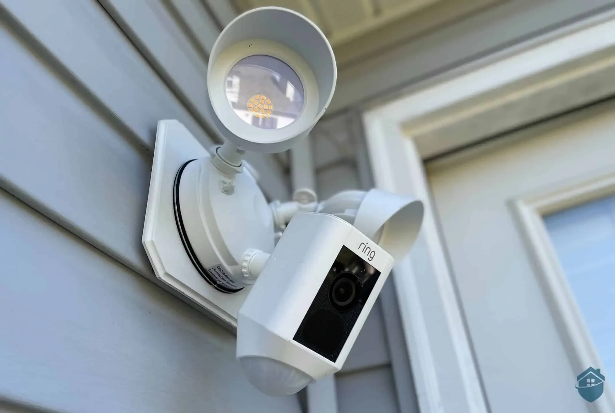 Ring security camera mounted on a house exterior wall with a white protective cover and a motion sensor light above it.