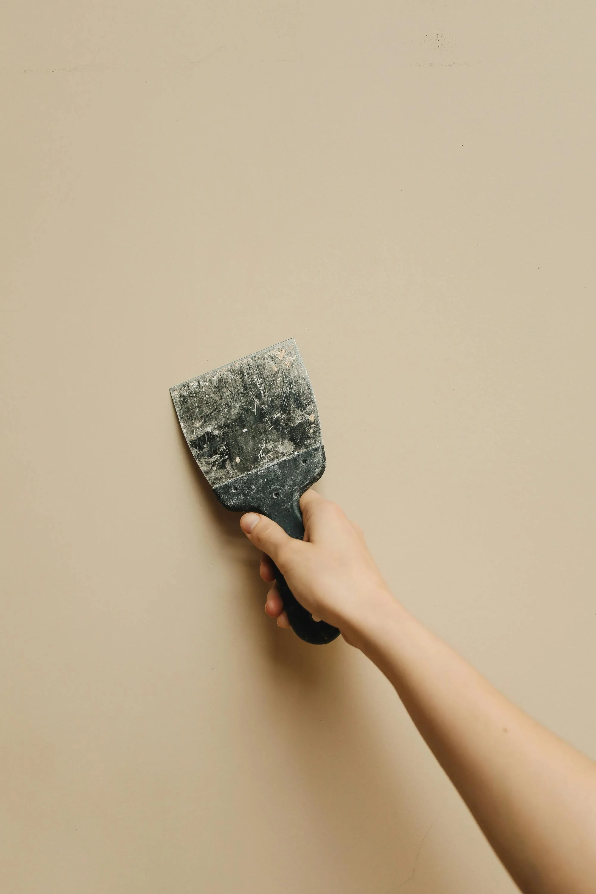 A person's hand holding a putty knife on a beige wall during drywall repair.