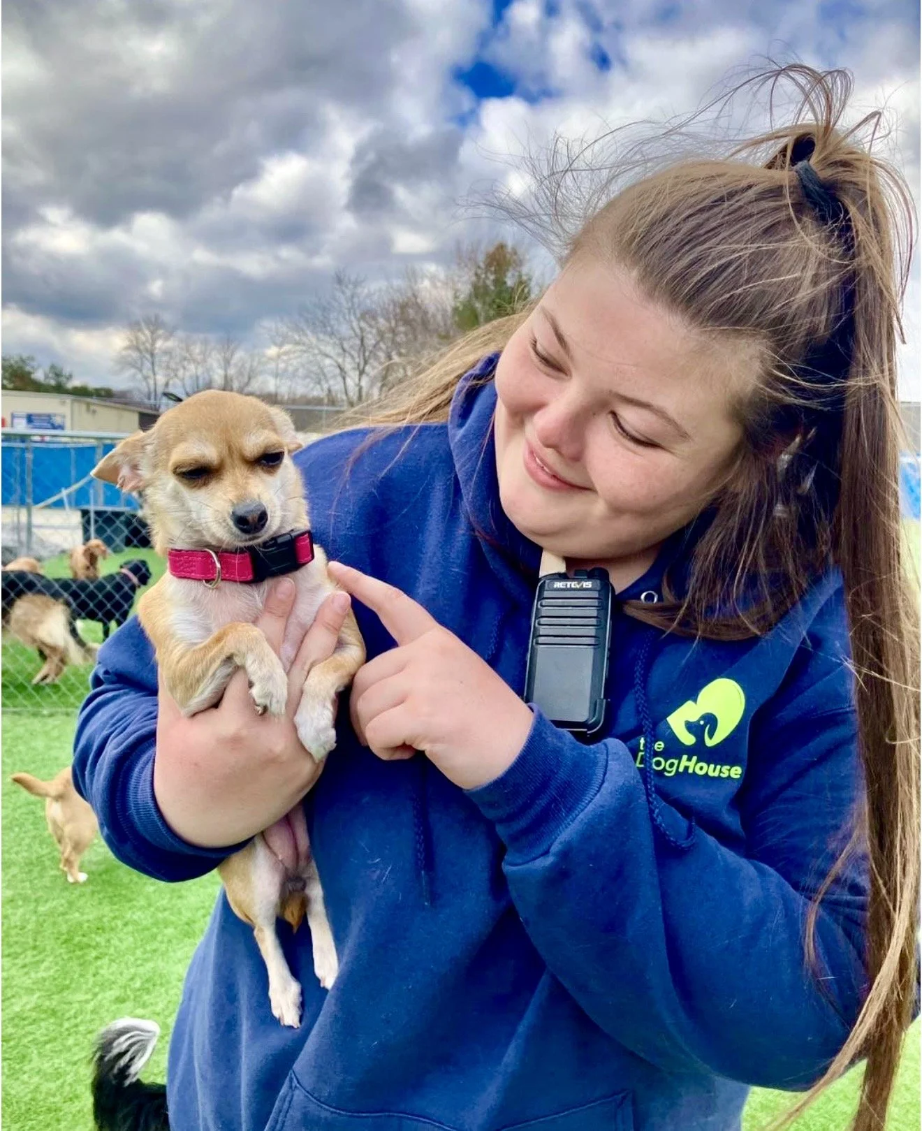 A woman holding a small brown and white dog with a red collar, smiling and touching its nose outside at a dog rescue or kennel, with a chain-link fence and other dogs in the background.