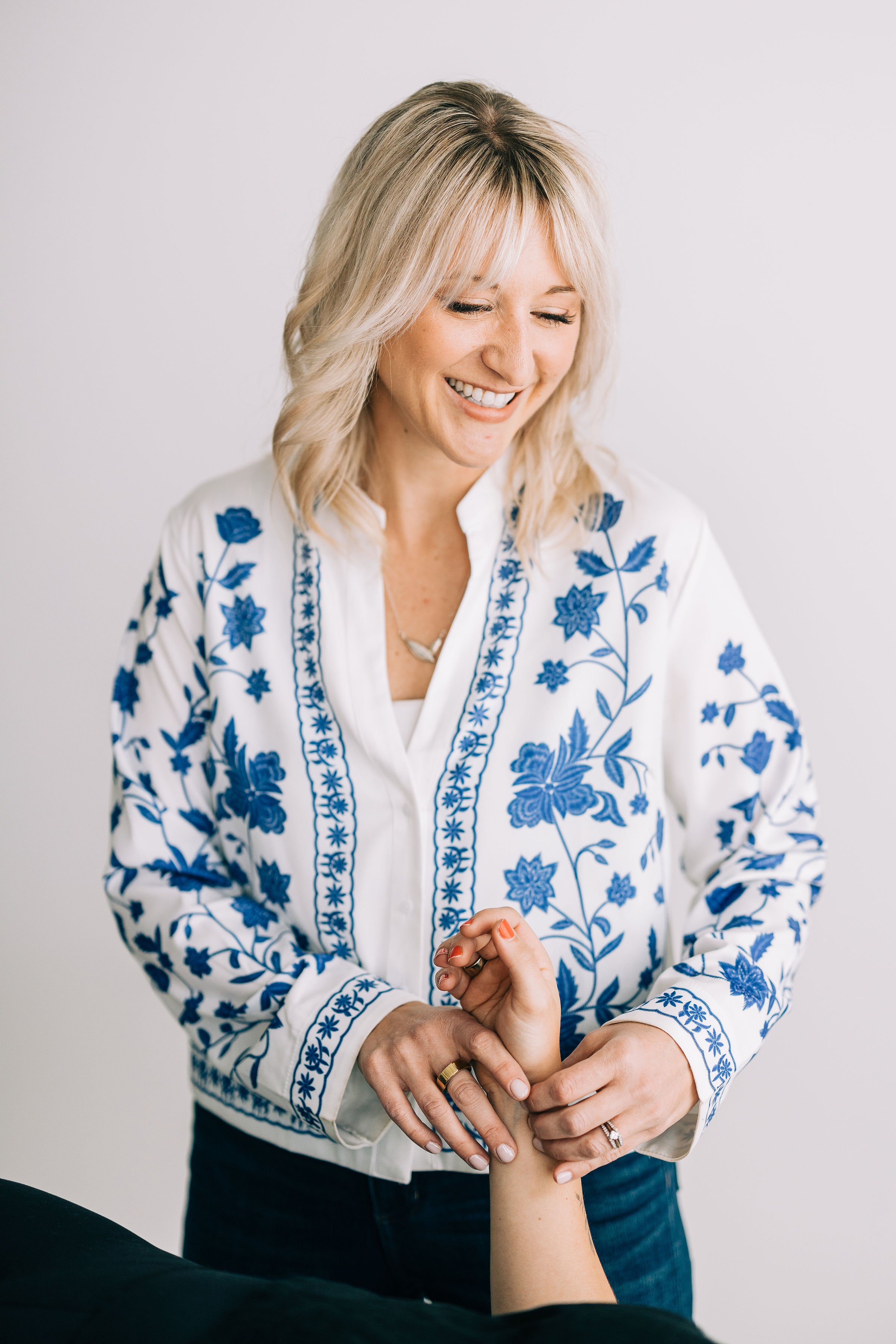 A woman with blonde hair smiling and looking down while receiving physical therapy on her arm from a woman wearing a white shirt with blue embroidery.