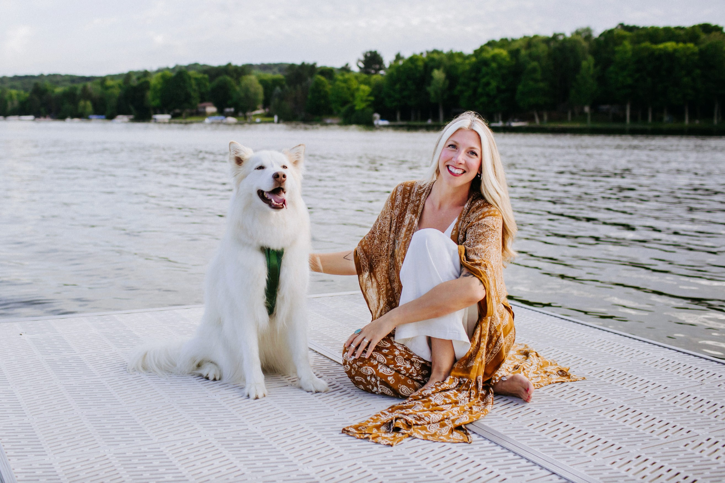 A woman with long blonde hair, smiling, sitting on a dock by a lake with a white dog sitting beside her. The woman is wearing a patterned dress and the dog has a green leash. The background shows trees and houses across the water.