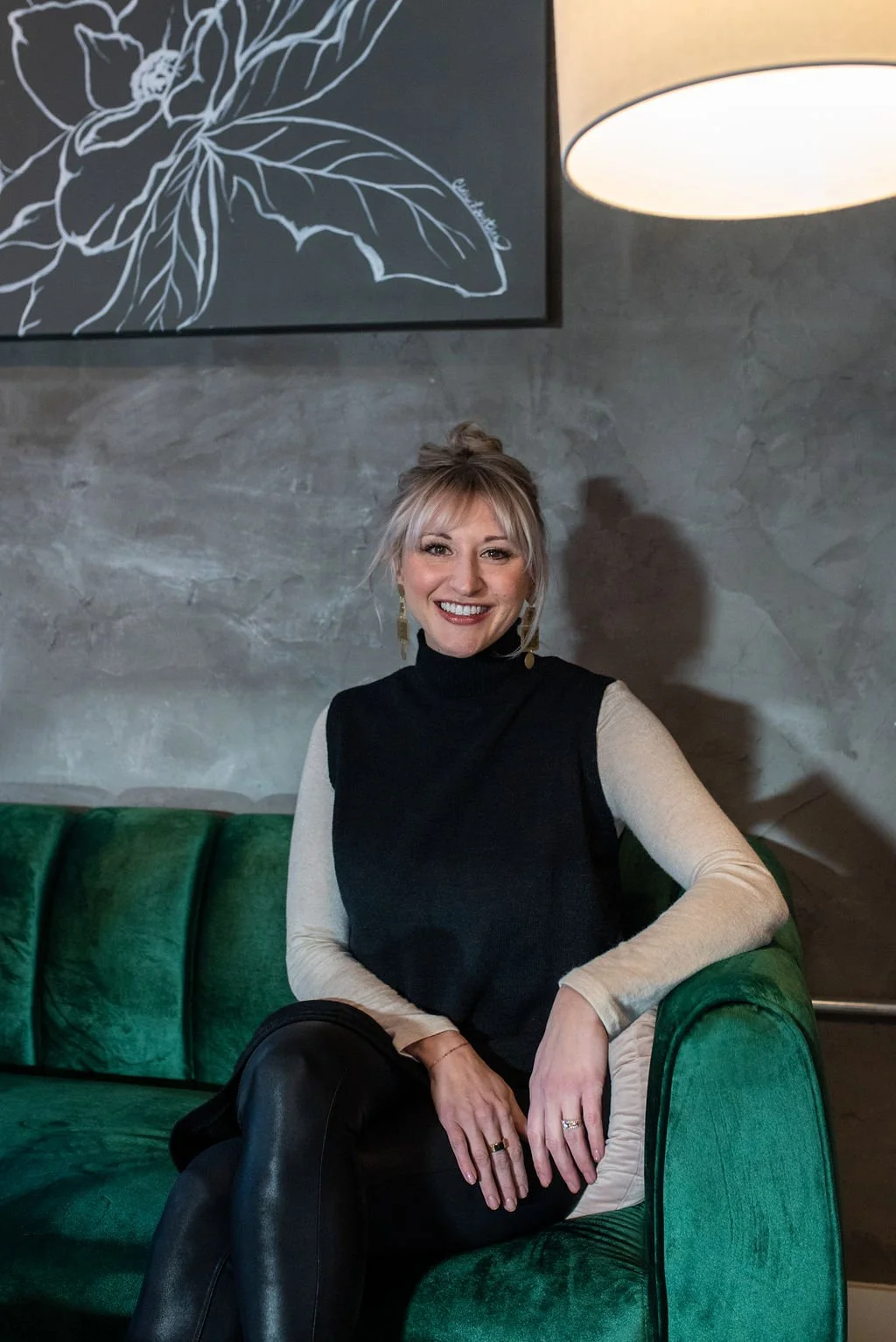A woman sitting on a green velvet sofa in a modern interior with a textured gray wall, a large leaf artwork, and a white hanging lamp