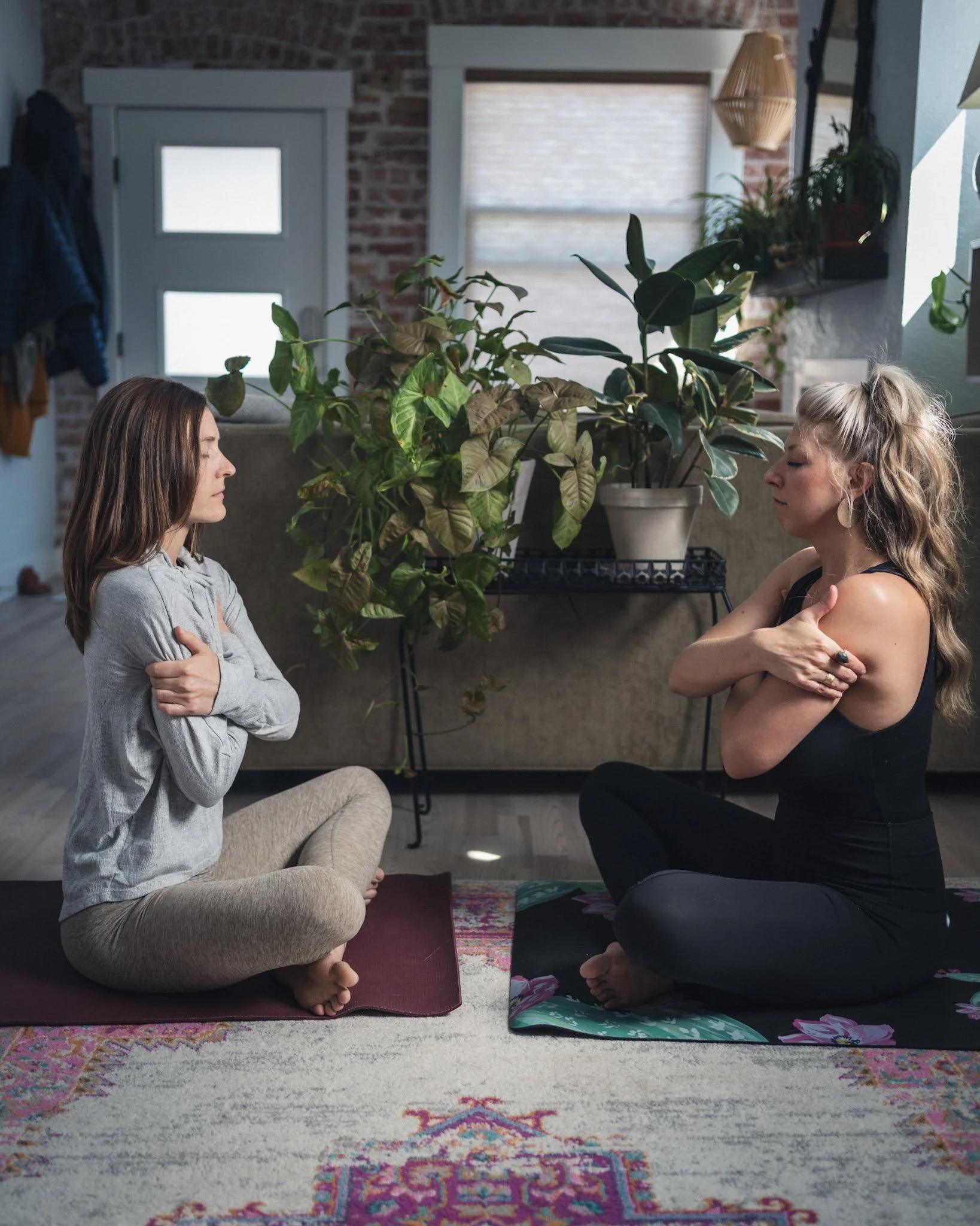 Two women practicing meditation or mindfulness in a cozy living room with indoor plants, one sitting cross-legged on a purple yoga mat and the other in a kneeling position on a patterned rug, both with eyes closed and arms crossed over their chests.