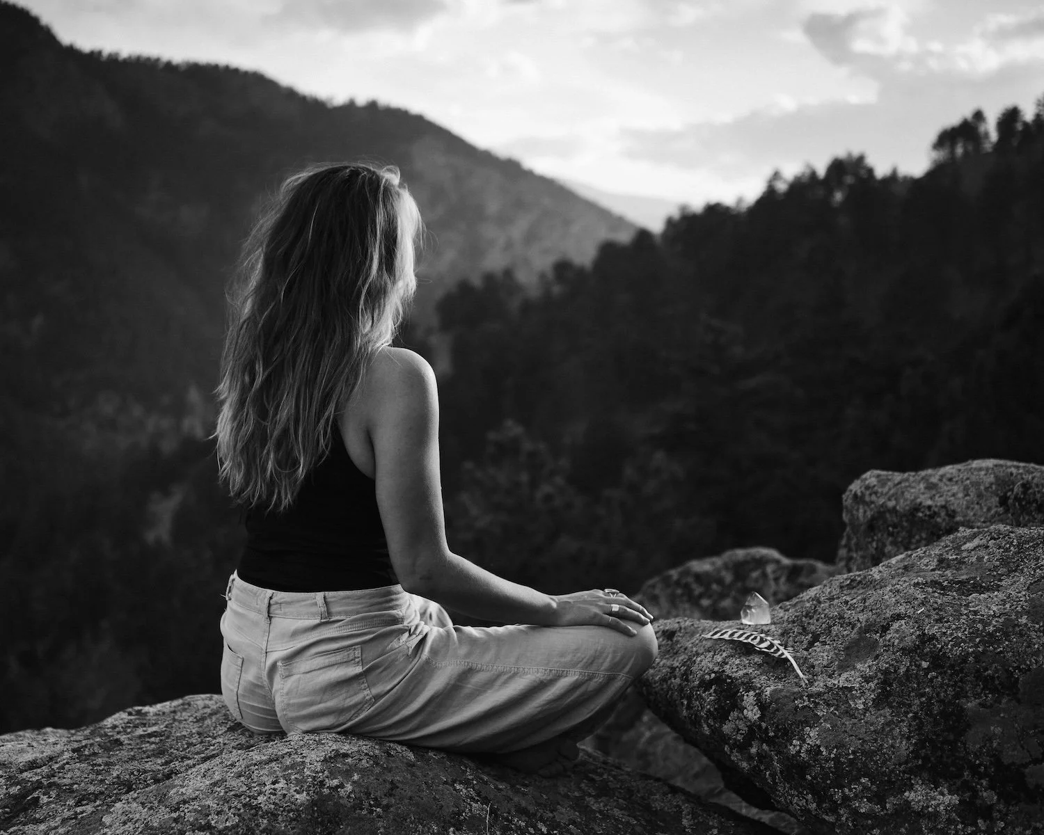 A woman with long hair sitting cross-legged on a rock, facing away, overlooking mountains in the distance, in black and white.