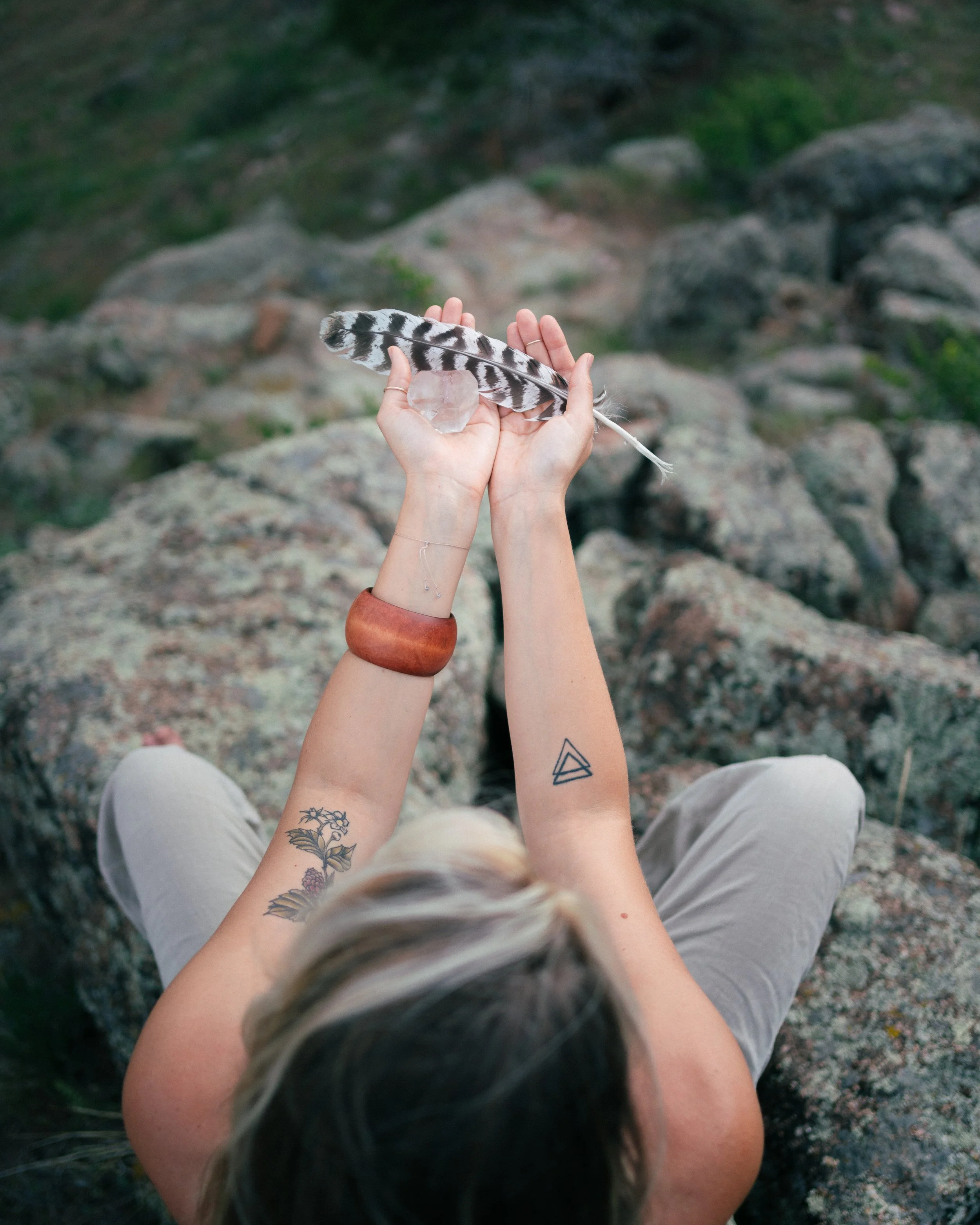 Person holding a black and white striped feather with a small clear crystal in the background, sitting on rocks in an outdoor setting.