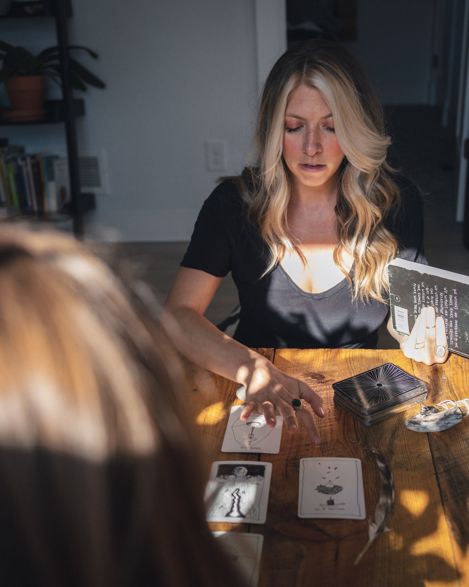 A woman with long blonde hair and a black shirt is sitting at a wooden table, looking down and organizing tarot or oracle cards. There are some cards laid out in front of her, including one with a chalice symbol. A deck of cards and a booklet or instruction sheet are also visible on the table. In the background, a bookshelf and some shelves with books and a potted plant can be seen.