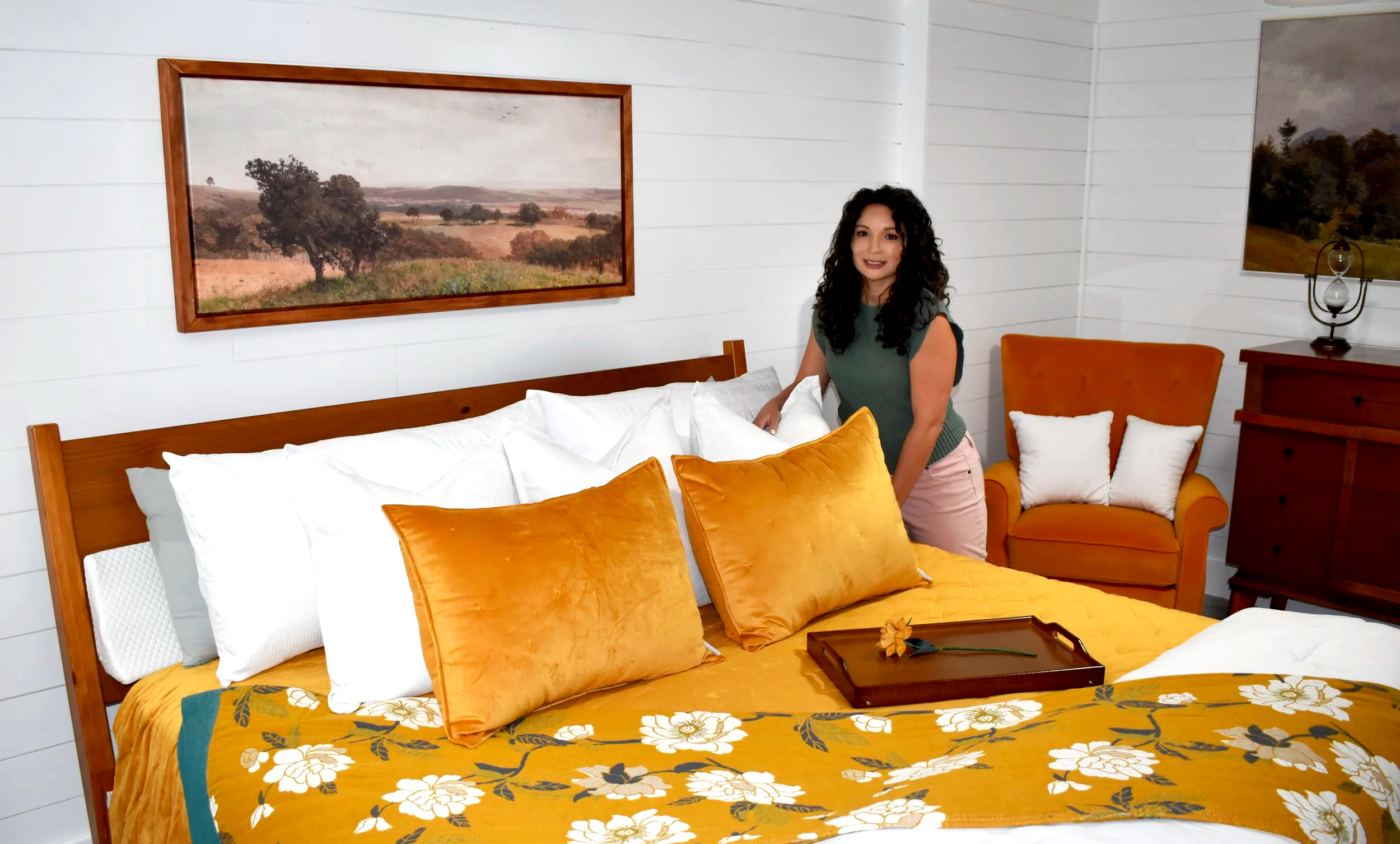 A woman with curly dark hair standing beside a bed in a room with white shiplap walls. The bed has white and yellow pillows and a floral yellow bedspread. There is a wooden nightstand, an orange armchair with white pillows, and framed landscape paintings on the walls.