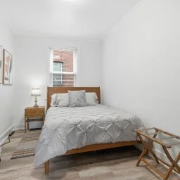 Bedroom with white walls, wooden bed frame, gray bedding, a small wooden nightstand with a lamp, and a window showing a brick building outside.