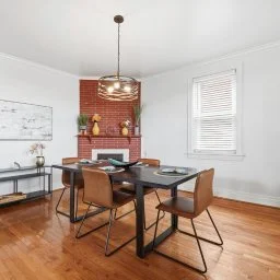Dining room with a black table and six brown chairs, brick fireplace with decor and books, white walls, window with blinds, hardwood floor, and a modern pendant light.