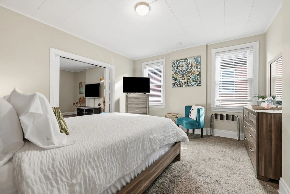 A bedroom with a large bed, dresser, and a chair, with windows and a television.