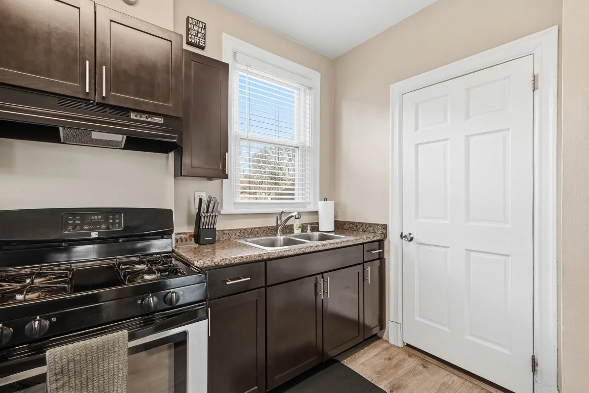 Kitchen with dark brown cabinets, a black stove, stainless steel sink, granite countertop, window with blinds, paper towel roll, and a white door.