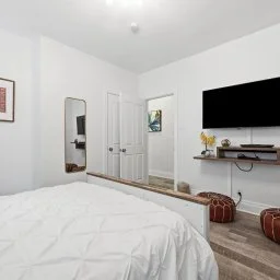 A bedroom with white walls, a bed with white bedding, a wall-mounted flat-screen TV, two brown poufs, a mirror, a small window, and a door.