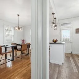 A view of a dining area and kitchen separated by a wall. The dining area has a wooden table with three chairs and a hanging light fixture. The kitchen has white cabinets, a countertop with fruit, and a window with blinds.
