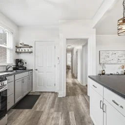 Bright, modern kitchen with white cabinets, black countertops, a window with blinds, and a hallway leading to other rooms.